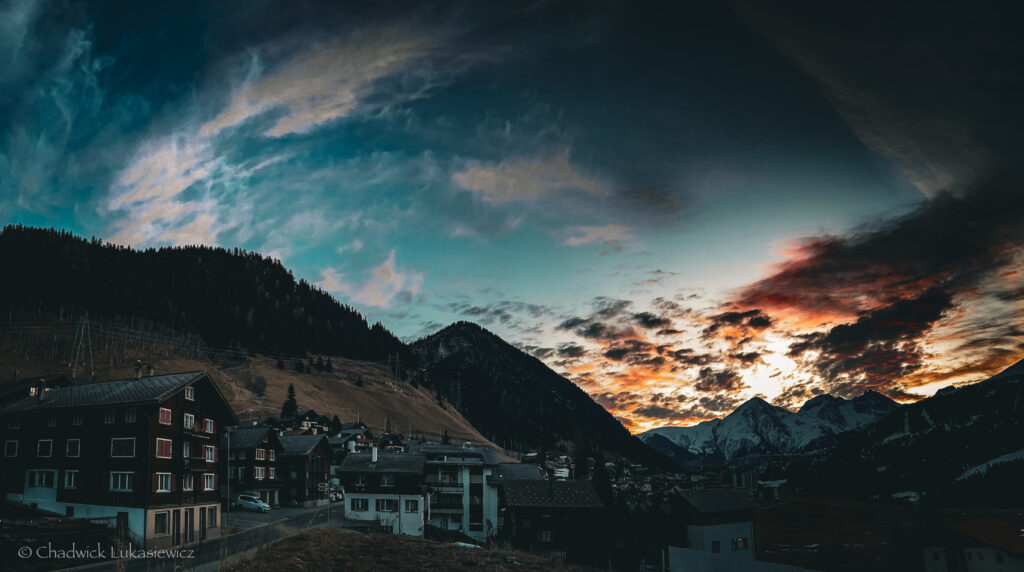 A panoramic view of a Swiss mountain village at sunrise, with snow-capped peaks in the distance and dark clouds glowing with hues of orange and pink as the sun rises. The village below is nestled between steep hills, with traditional alpine homes, and a soft light touches the tops of buildings and mountains. The sky transitions from deep blue to vibrant sunrise colors, casting a peaceful glow over the scene.