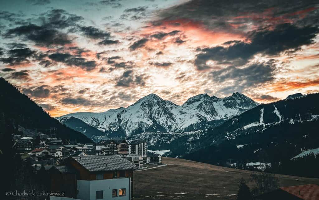 Snow-capped mountains in Sedrun, Switzerland, illuminated by a vibrant sunset sky, with a small village in the foreground and patches of snow on the surrounding hills.