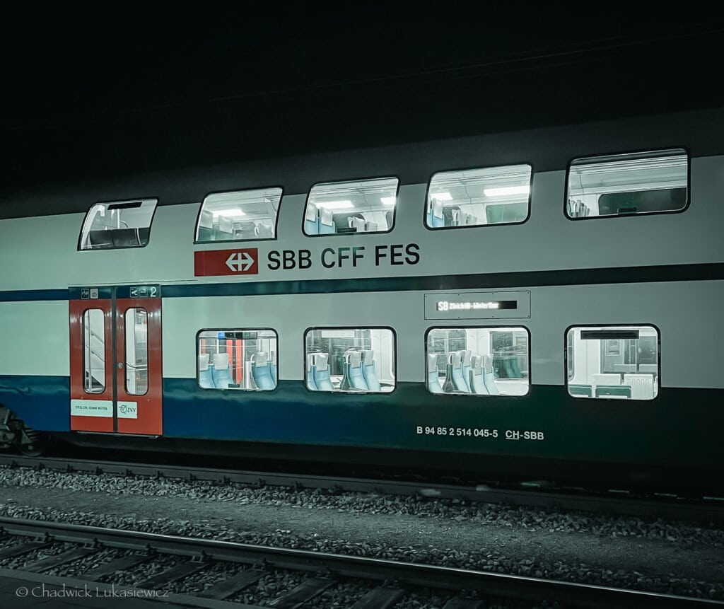 A double-decker SBB CFF FES train parked on a track at night. The train’s well-lit interior is visible through large windows, showcasing empty rows of seats. The red and white exterior stands out against the dark surroundings, with the station’s dim lights reflecting softly on the train.