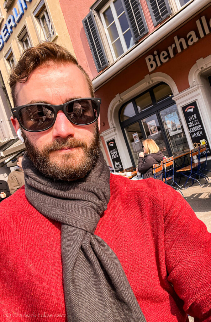 Self-portrait of a bearded man wearing sunglasses, a dark scarf, and a bright red sweater, standing in front of a European-style building with “Bierhall” signage in Zurich, Switzerland. Outdoor seating with people enjoying food and drinks is visible behind him on a sunny winter day.