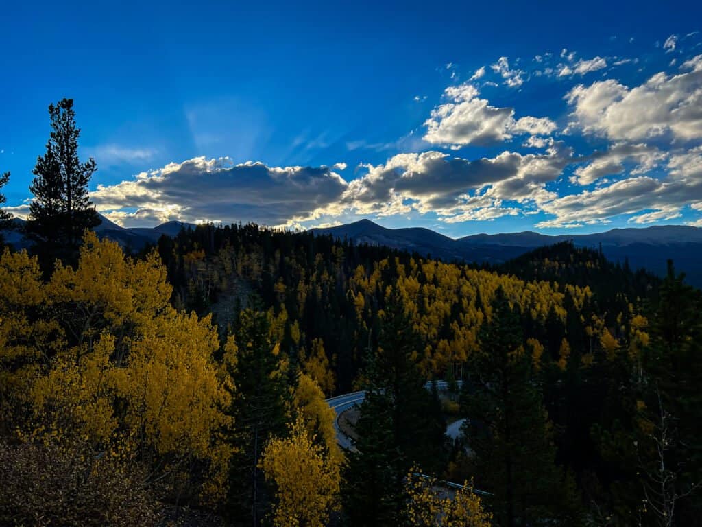 Golden autumn trees line a forested mountain landscape at sunset along Aspen Alley Trail in Breckenridge, Colorado, with clouds glowing above distant peaks.