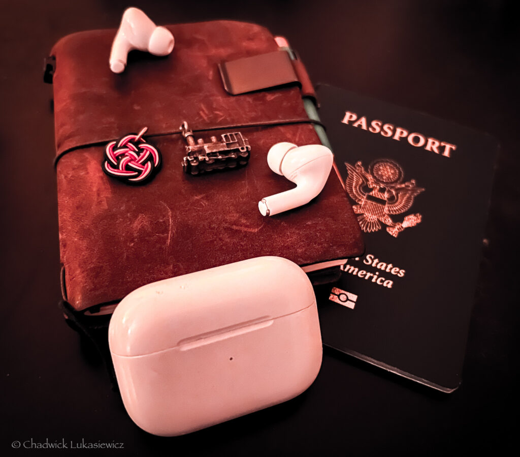 A pair of Apple AirPods Pro earbuds with their white charging case rest on top of a worn brown leather traveler’s notebook. The notebook is adorned with a small red knot decoration, a tiny metal train charm, and a black pen clipped to it. A U.S. passport with its dark blue cover is partially visible beneath the notebook, all set against a dark background.