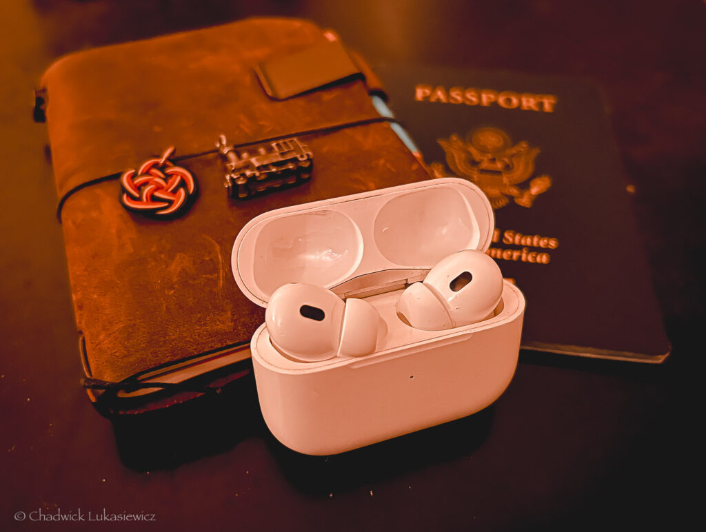 A white AirPods Pro case with the earbuds resting open on a dark surface, accompanied by a worn leather traveler’s notebook adorned with a train charm and a red knot, and a U.S. passport in the background.