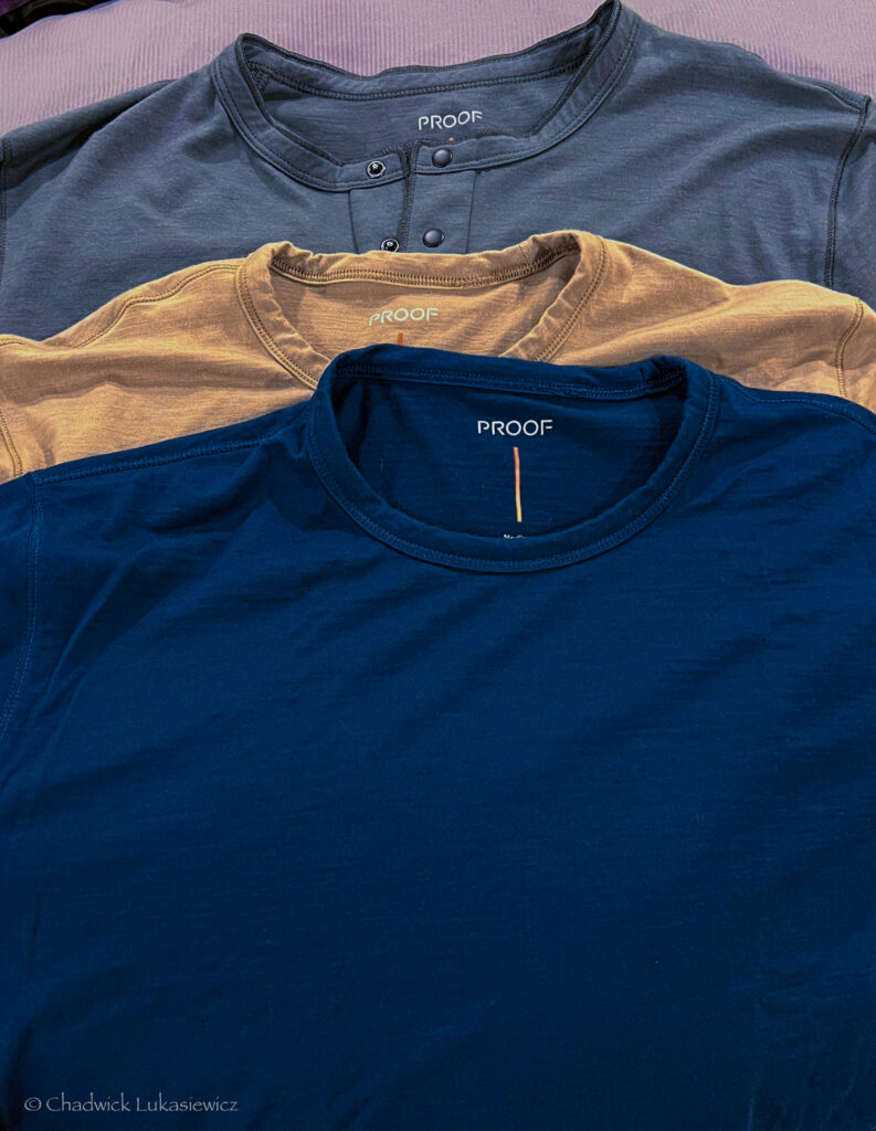 A neatly arranged display of three “PROOF” branded shirts, stacked one on top of the other. The top shirt is a dark gray henley with two buttons near the collar. The middle shirt is a light tan color, and the bottom shirt is a rich navy blue. All shirts are part of the Huckberry “72-Hour” collection, as seen from the visible tags. The soft texture and casual style suggest they are made from comfortable, high-quality fabric