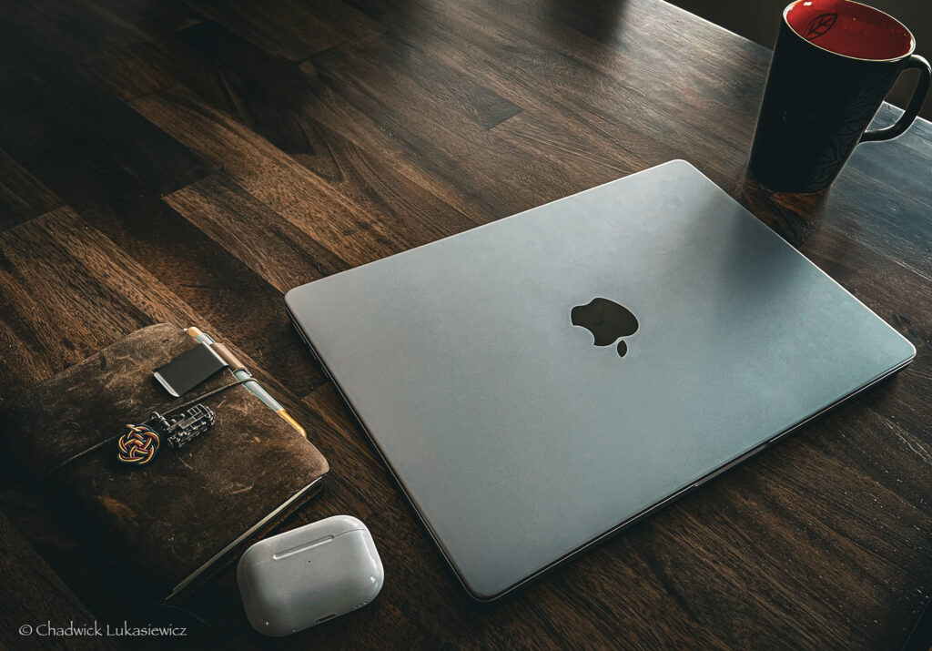 A neatly arranged workspace featuring a closed silver MacBook, a leather-bound notebook adorned with a train-themed charm, a pen tucked into the notebook’s elastic band, and a pair of Apple AirPods resting nearby. A black ceramic coffee mug with a red interior sits on the wooden desk, completing the minimalist setup for remote work or journaling. The wooden surface adds warmth to the scene, suggesting a calm and focused environment.