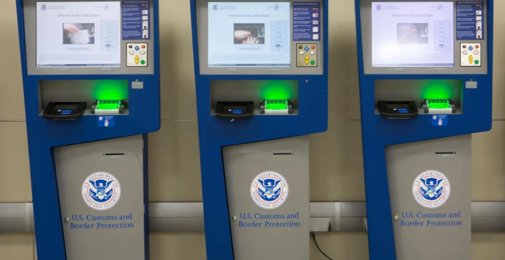 Three blue Global Entry kiosks used by U.S. Customs and Border Protection are aligned next to each other. Each kiosk has a touchscreen display at the top with instructions, a passport scanner, and a fingerprint scanner that glows green. The U.S. Department of Homeland Security emblem is prominently displayed on the lower section of the machines. These kiosks are designed to expedite the customs process for pre-approved travelers returning to the United States.