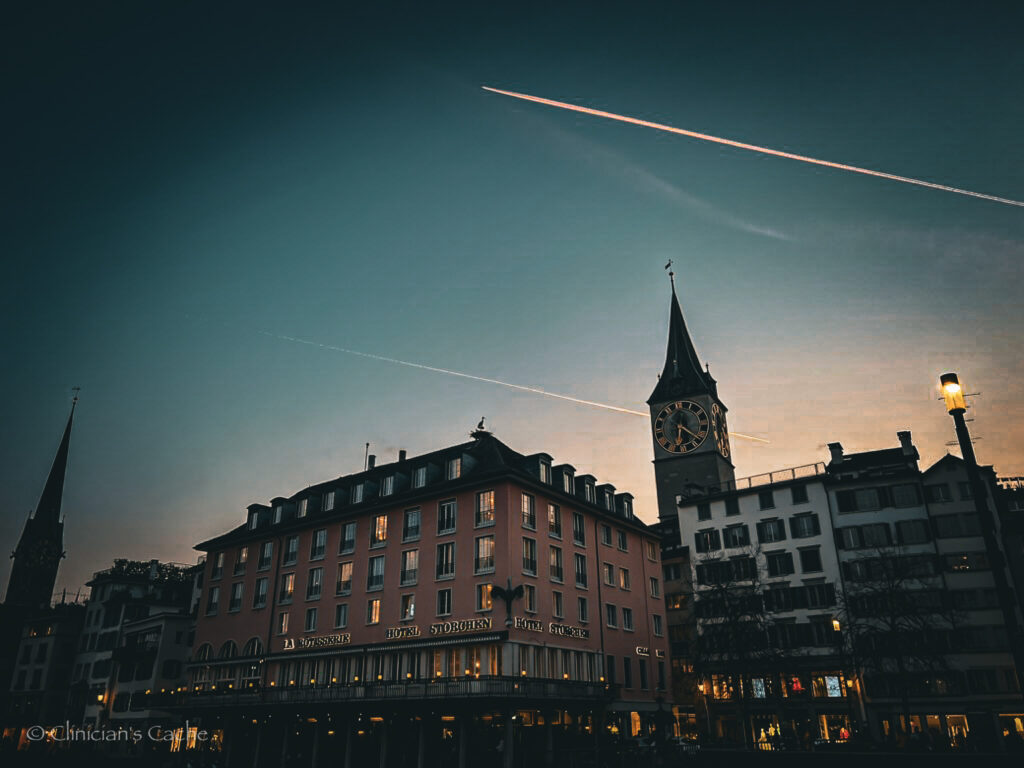 Evening view of a clock tower and buildings in Zurich, Switzerland, with airplane contrails crossing the sky and warm lights beginning to illuminate the street.