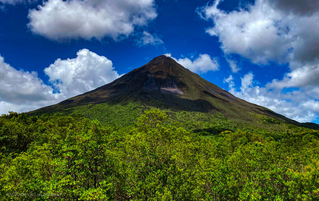 A clear view of Arenal Volcano in Costa Rica, towering above a lush green forest under a bright blue sky dotted with white clouds. The volcano’s steep cone is prominent, with visible patches of darker rock contrasting against its green base.