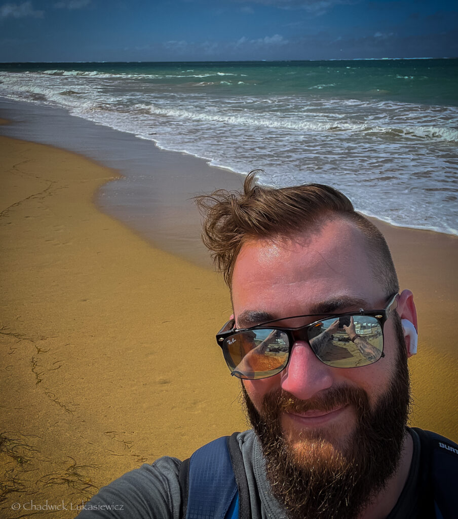 Self-portrait of a man with a beard, sunglasses reflecting the beach surroundings in Puerto Rico, and Apple AirPods Pro in his ears, standing on a sunny golden beach with ocean waves gently washing ashore. The clear blue sky and turquoise water extend into the distance.