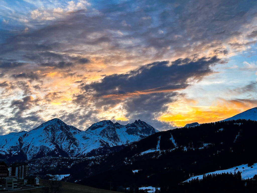 Snow-capped mountain range in Sedrun, Switzerland, silhouetted against a vibrant sunset sky with dramatic clouds.