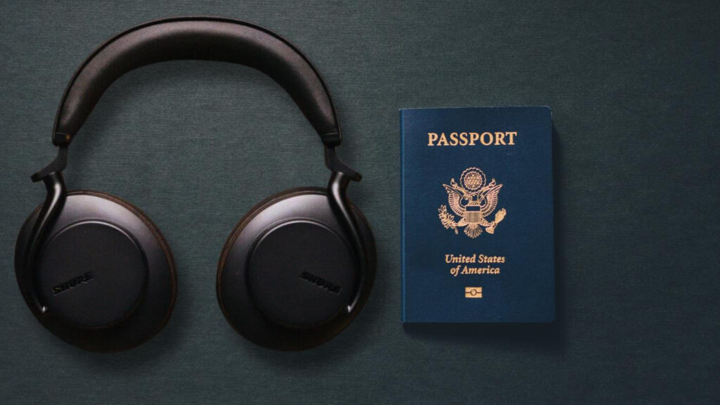 A pair of black Shure AONIC 50 Gen 2 headphones lying flat on a dark green textured surface, with a blue United States passport positioned to the right. The headphones have a sleek, modern design with round ear cups and a padded headband. The passport cover features the gold embossed seal of the United States