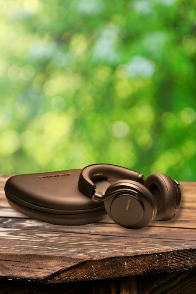 A pair of black Shure AONIC 50 Gen 2 headphones resting on a matching hard-shell case, placed on a rustic wooden table outdoors. The blurred background features lush green foliage, creating a vibrant, natural setting.