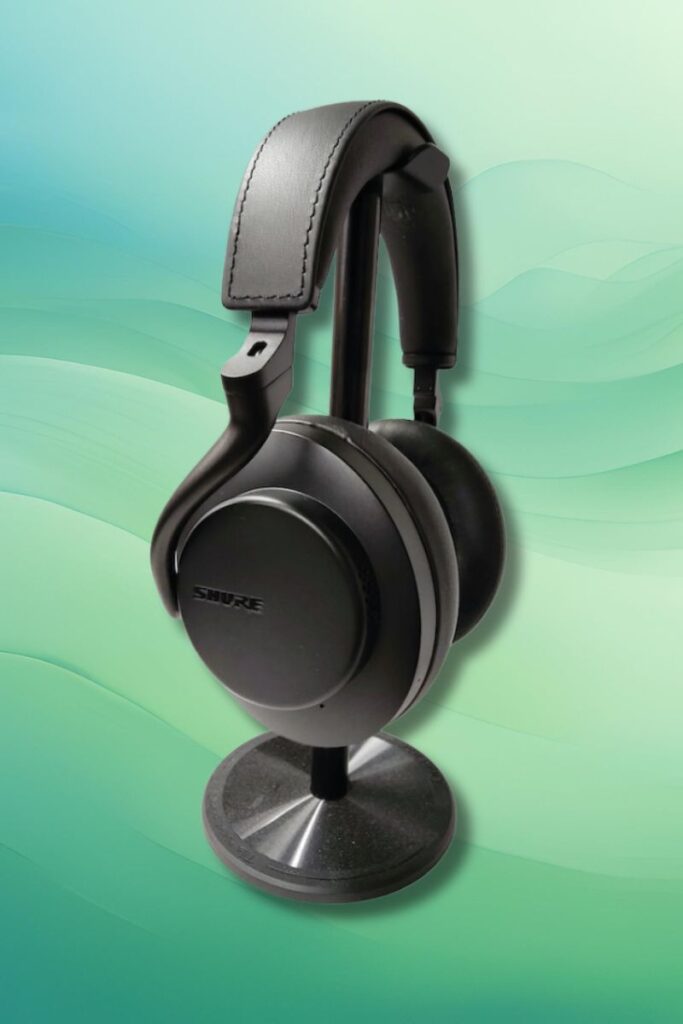 A pair of black Shure AONIC 50 Gen 2 over-ear headphones displayed upright on a metal headphone stand with a circular base against a soft green gradient background. The ear cups are round with the “Shure” logo prominently visible.