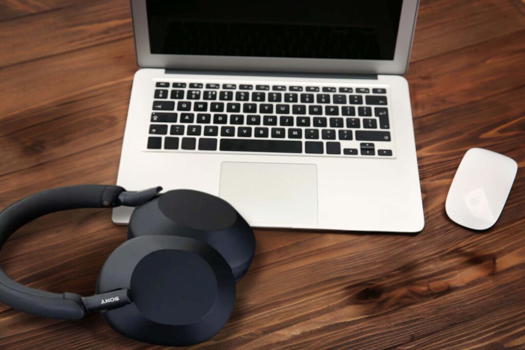 A pair of black Sony WH-1000XM5 over-ear headphones resting on a wooden table next to a silver laptop with a black keyboard and an Apple Magic Mouse.