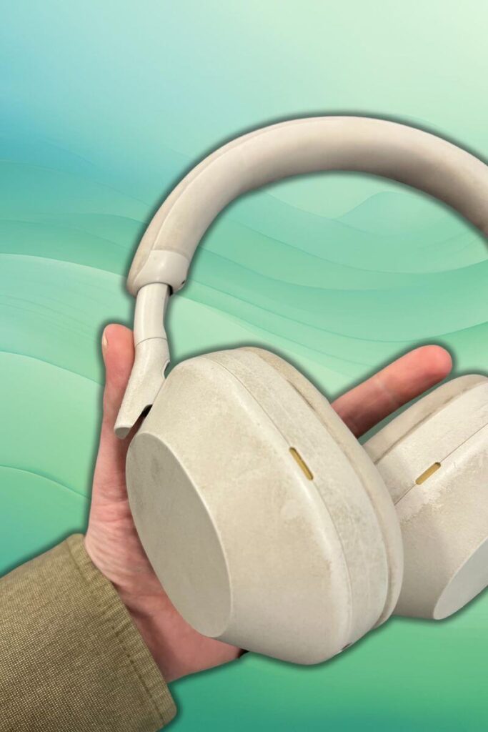 A pair of white Sony WH-1000XM5 over-ear headphones, appearing slightly dirty and worn, held in hand. The earcups and headband are off-white, set against a gradient green background.