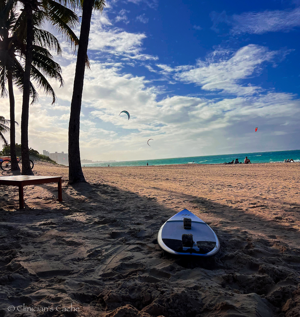 A surfboard resting on the sandy beach under palm trees in Puerto Rico, with kite surfers in the distance and a bright blue sky filled with clouds