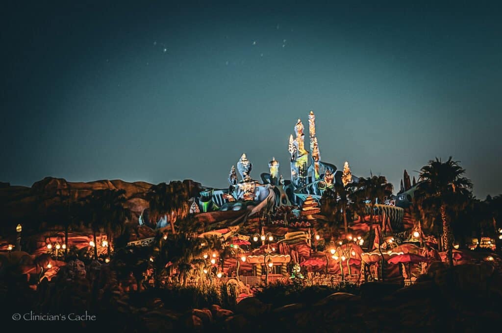 An illuminated fantasy-themed building resembling an underwater palace, glowing against the night sky, surrounded by vibrant lights and palm trees in Tokyo, Japan.