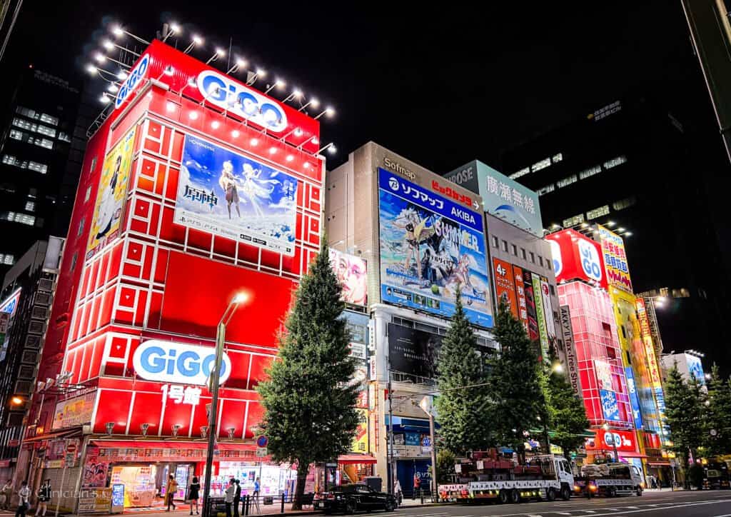 A vibrant night scene in Tokyo’s Akihabara district, featuring brightly lit buildings with colorful billboards, including large advertisements for games and anime on the red GiGO arcade building. Tall trees line the street below, with cars and pedestrians in view.