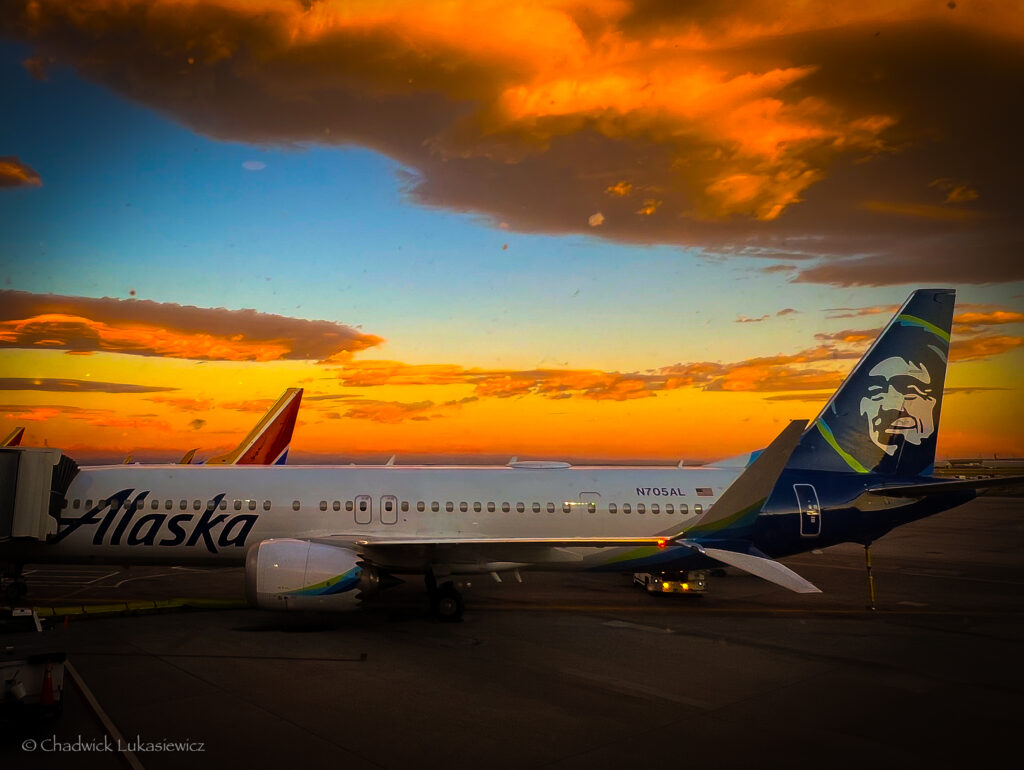 A side view of an Alaska Airlines plane at Denver International Airport during sunrise. The plane’s tail features the distinctive face logo of the airline, while the sky above is ablaze with dramatic orange and yellow clouds. Another plane’s tail can be seen in the background, and the airport tarmac is visible below. The warm glow of the rising sun creates a striking contrast against the cool tones of the aircraft.