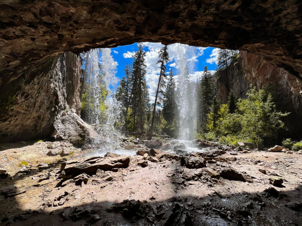 A stunning view from behind a waterfall at Hanging Lake near Glenwood Springs, Colorado. Water cascades down from the rocky overhang, with sunlight filtering through, casting shadows on the damp stones. Tall pine trees and lush greenery surround the area, with a bright blue sky filled with fluffy clouds visible in the background. The natural beauty of the scene is accentuated by the dynamic contrast of the rocky cave and the flowing water.