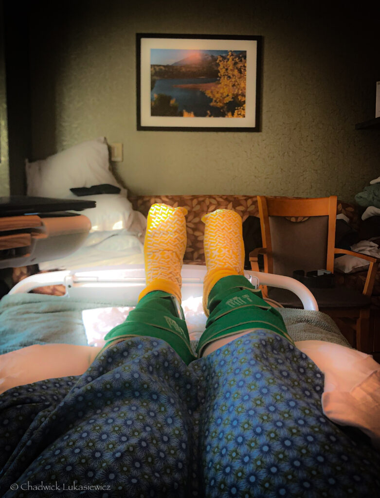 A person is lying in a hospital bed, with their legs extended and covered by patterned hospital blankets. They are wearing bright yellow non-slip socks, and green medical compression devices are wrapped around their lower legs. In the background, a framed landscape photo hangs on the wall, depicting a serene scene of a lake and mountains. The room is dimly lit, giving the image a calm and quiet atmosphere