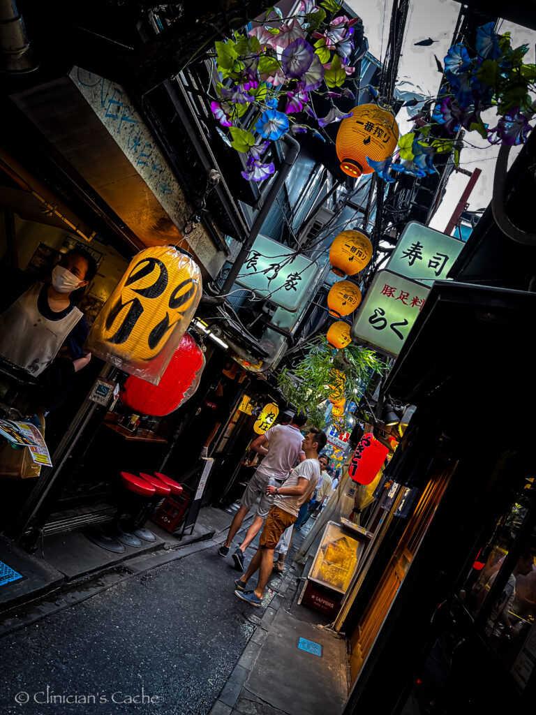 Narrow street market in Tokyo, Japan, lined with colorful paper lanterns and signs, as people walk past small restaurants and shops.