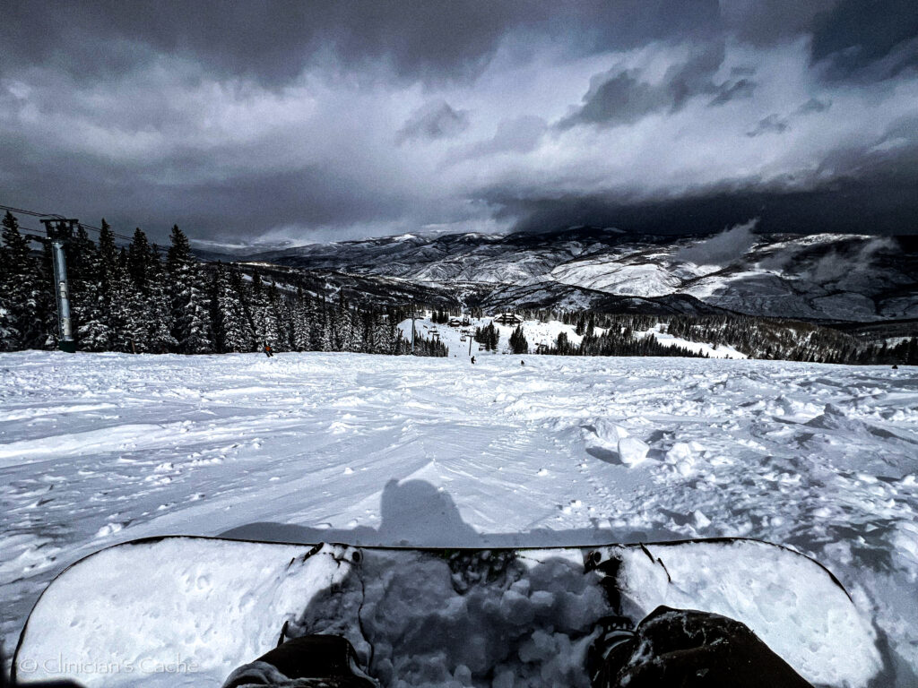 Snowboarder’s perspective sitting on a snowy slope at Beaver Creek, Colorado, surrounded by pine trees and overlooking snow-covered mountains under a cloudy sky.