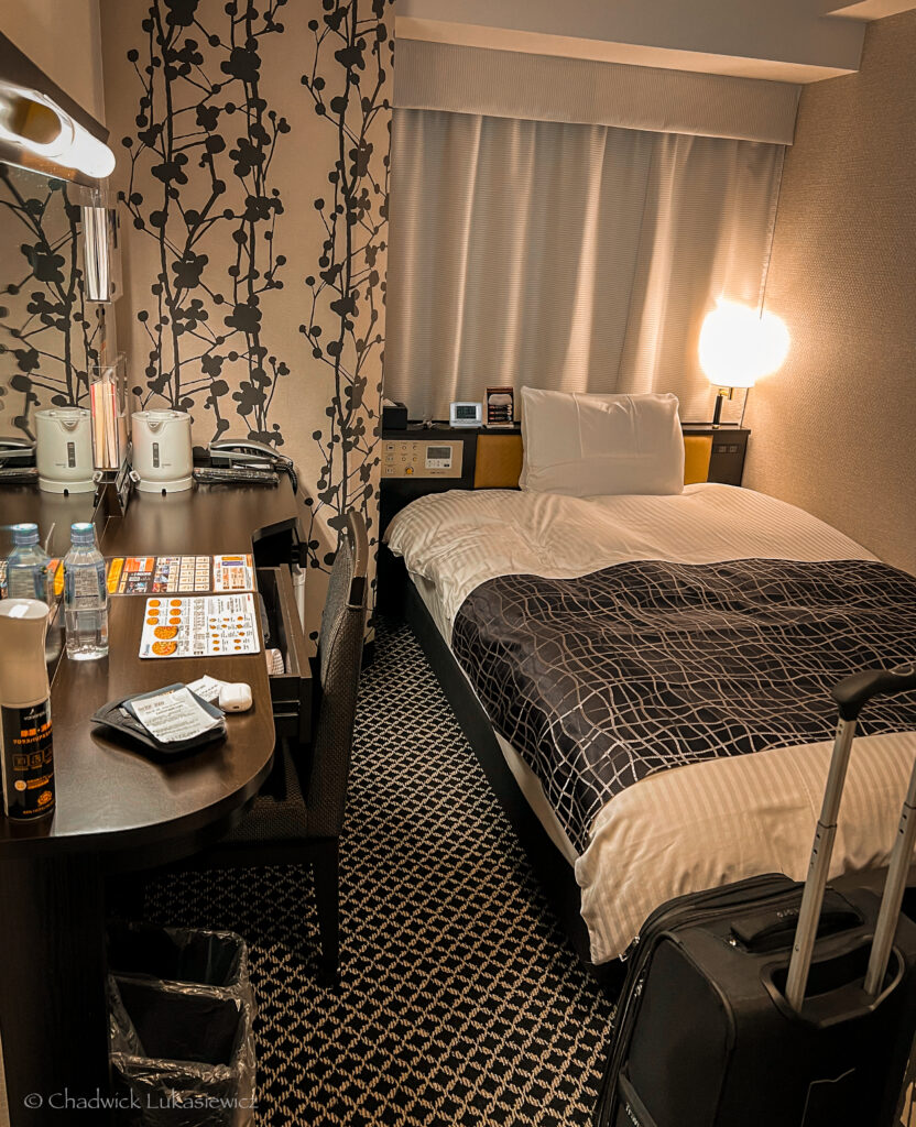 compact Japanese hotel room with a single bed dressed in black and white linens. A small desk equipped with a kettle, remote controls, and complimentary water bottles sits nearby. The room’s walls feature a patterned floral design, and a suitcase stands near the bed, reflecting a cozy, minimalist setup typical of Japanese business hotels.