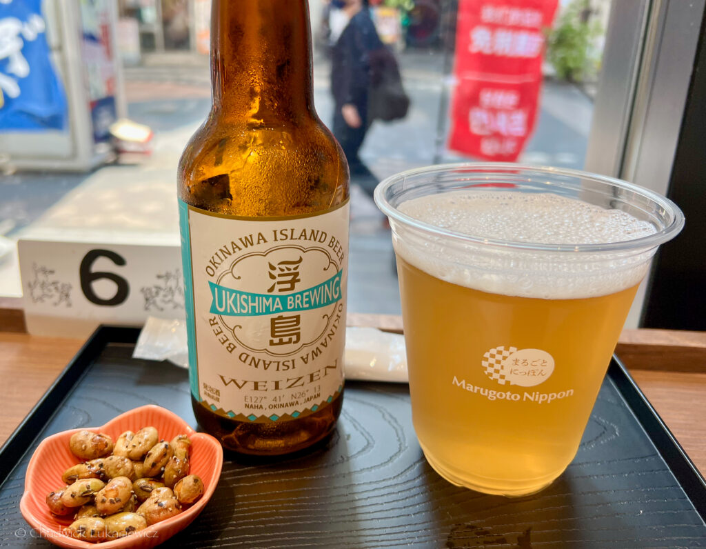 A glass of Ukishima Brewing’s Okinawa Island Weizen beer sits on a black tray beside its bottle, displaying Japanese branding. The tray also includes a small pink dish filled with seasoned nuts. A table marker with the number six is visible in the background, with street activity slightly blurred through the nearby window.