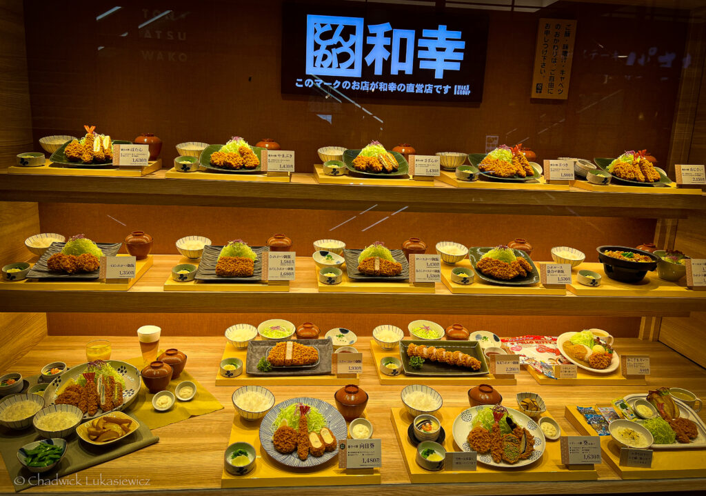 A glass display showcasing various plated tonkatsu meals from a Japanese restaurant. Each meal is arranged with garnishes, sides, and prices listed in Japanese yen. The neatly organized rows highlight the variety of deep-fried pork cutlet dishes, accompanied by rice, soup, and small bowls of pickled vegetables.