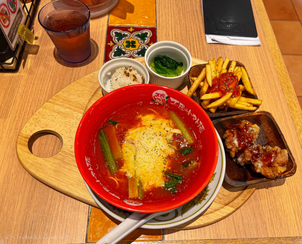  A colorful Japanese meal centered around a red bowl of tomato-based ramen topped with vegetables and cheese. The meal includes side dishes such as rice, seasoned greens, French fries with tomato sauce, and two pieces of fried chicken, all served on a wooden tray. A glass of water and a decorative table setting complete the presentation