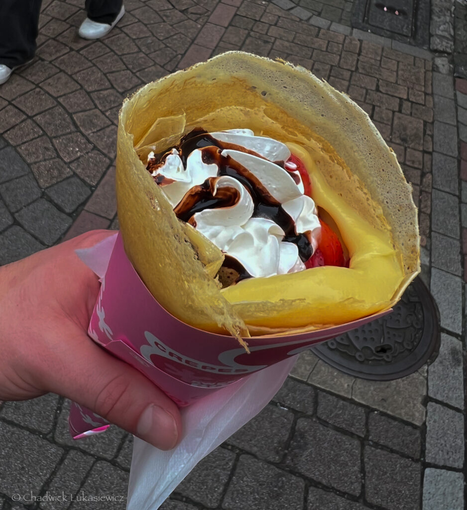 A close-up of a hand holding a Japanese-style crepe from a street vendor in Harajuku, Tokyo. The crepe is wrapped in a pink paper cone and filled with whipped cream, chocolate drizzle, and visible fresh strawberries. The golden crepe edges are crisp and slightly folded over the toppings, and the background shows a textured cobblestone street