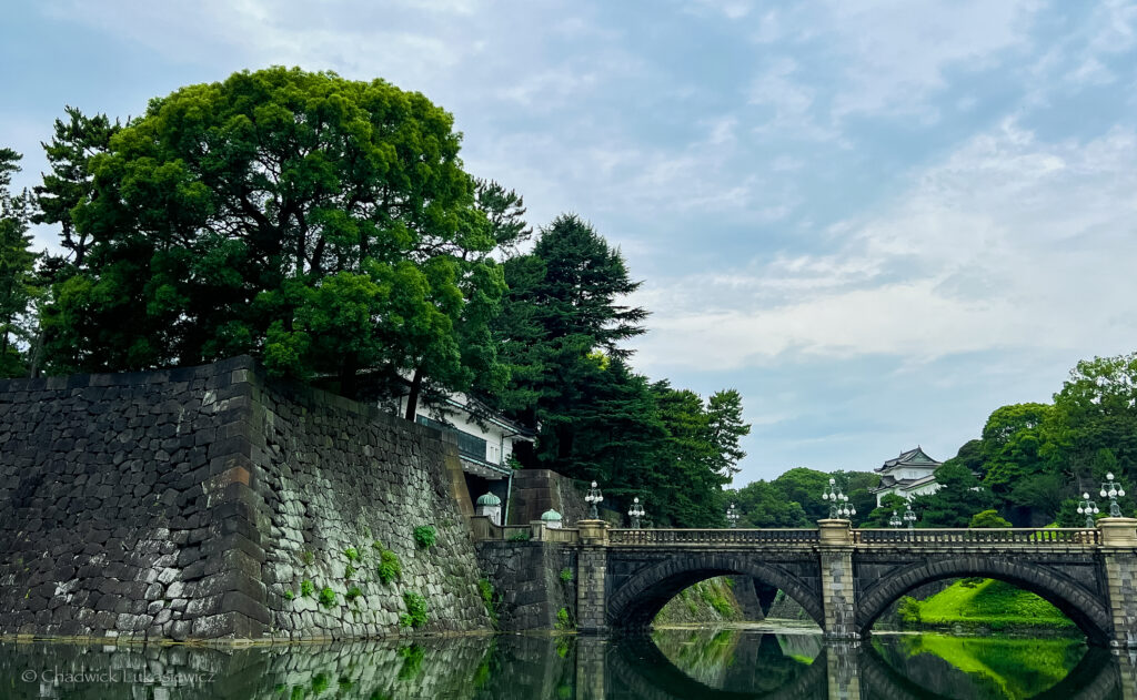 tranquil scene at the Kokyo Gaien National Garden in Tokyo, featuring a historic stone bridge arching over a still body of water, with its reflection visible on the surface. Large, lush trees surround the area, and a portion of the traditional Japanese Imperial Palace is seen in the background, partially obscured by greenery. The stone walls of the palace grounds rise along the left side, adding a sense of ancient architecture amid the peaceful natural setting. The sky above is slightly overcast, creating a calm atmosphere.