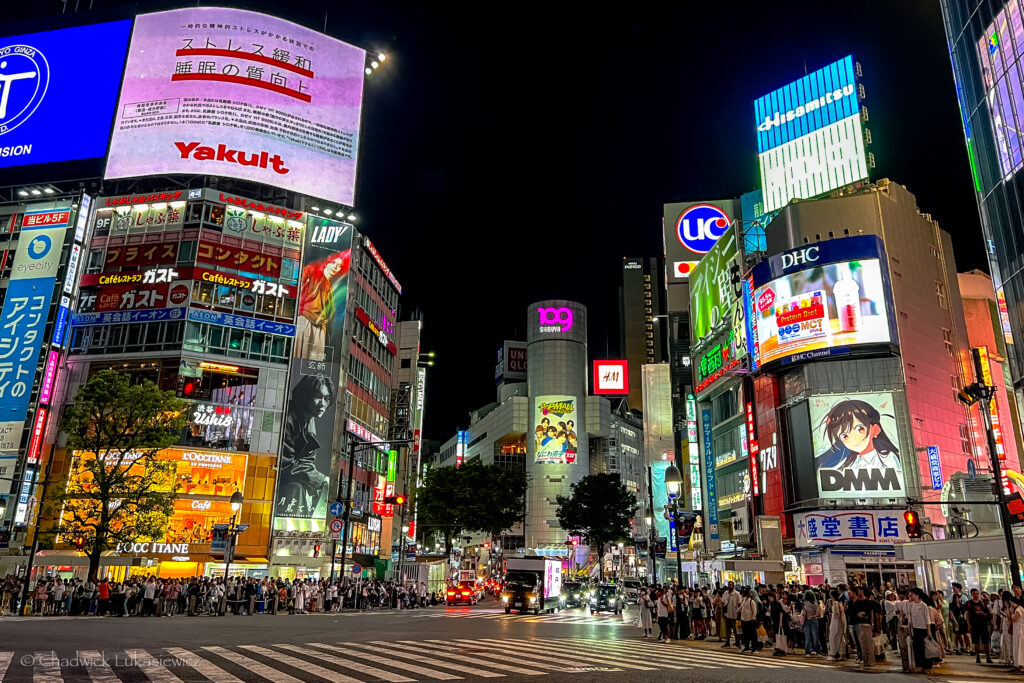 A bustling night scene at Shibuya Crossing in Tokyo, Japan, featuring large digital billboards and neon advertisements lighting up the surrounding buildings. Prominent signs include brands like Yakult, UC, and DHC, as well as the iconic Shibuya 109 shopping mall in the background. A crowd of people gathers along the sidewalks, waiting to cross the famous intersection. The street and buildings are vividly illuminated, capturing the vibrant energy of Tokyo’s nightlife in one of its busiest districts.