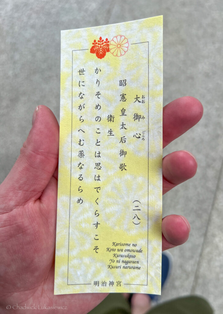 A hand holding a yellow card featuring Japanese text along with an English transliteration and translation. The text on the card is a waka poem from the Meiji Jingu shrine, encouraging tranquility and reflection. The intricate floral designs at the top add a decorative touch to the meaningful piece of paper.