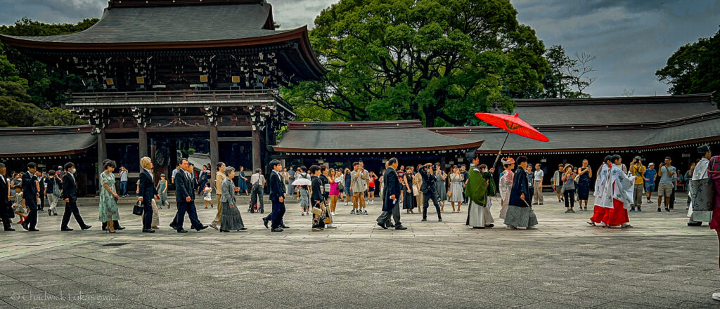 A traditional Japanese wedding procession at Meiji Jingu Shrine in Tokyo. The scene features a line of people dressed in formal attire, including traditional kimonos and formal suits, walking past the temple’s entrance. At the front, a person holds a large red parasol, a signature feature in Japanese wedding processions. The background shows the shrine’s ornate wooden gate, with its classic architectural style and surrounded by lush greenery. Spectators in casual attire gather around, watching and capturing the moment with cameras