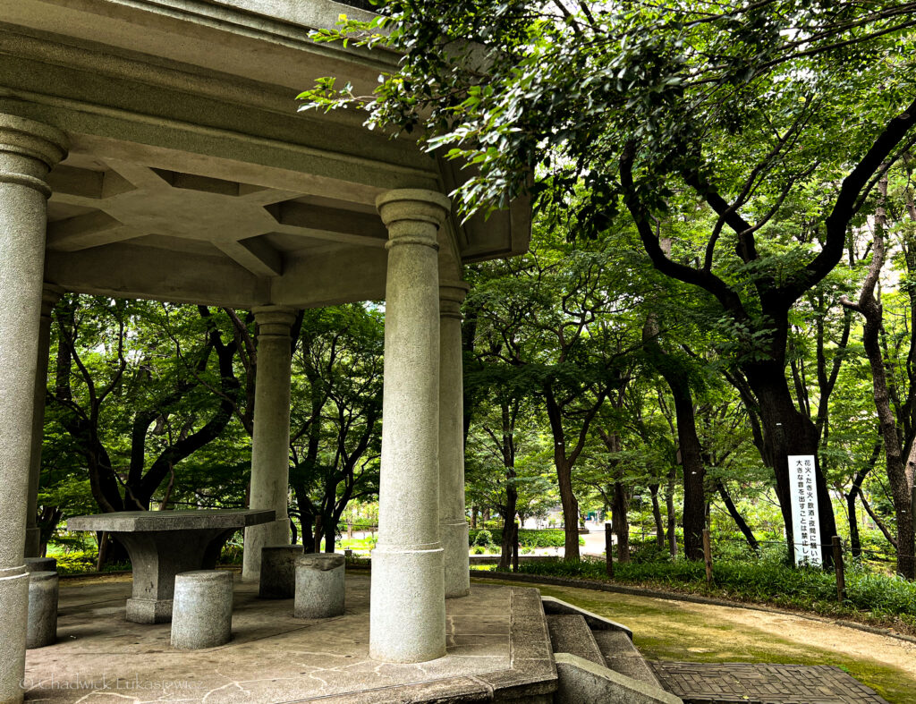 A stone pavilion in Shinjuku Chuo Park, Tokyo, featuring sturdy pillars and a central round table surrounded by cylindrical stools, all made of the same gray stone. The pavilion is nestled among dense greenery, with large trees creating a shaded, serene atmosphere. A path lined with lush vegetation winds around the pavilion, with a wooden signpost visible among the trees. Sunlight filters through the leaves, adding to the tranquil ambiance of this natural park setting.