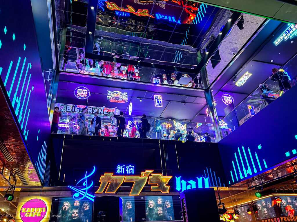 An interior view of a multi-level entertainment complex in Shinjuku, Tokyo, with vibrant neon signs and colorful lights. Crowds of people explore various levels, reflecting in mirrored surfaces above. Japanese signage and bright animations add to the lively, futuristic atmosphere.