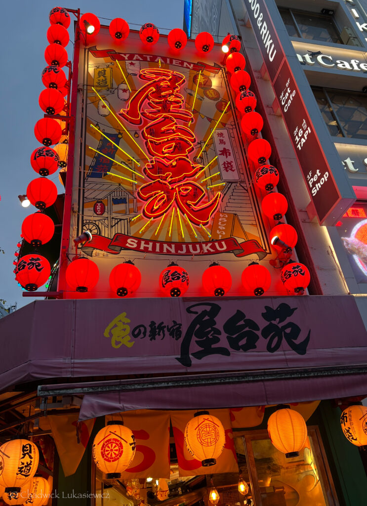 A bright, red neon sign with Japanese characters lights up the front of a restaurant in Shinjuku, Tokyo. The sign is decorated with red lanterns and reads “Shinjuku Yatai-en,” adding a vibrant pop of color against the evening sky, attracting passersby with its inviting glow.