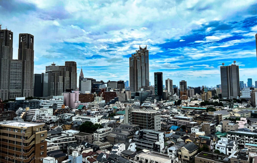 A wide-angle view of Tokyo’s skyline with a mix of modern high-rises and smaller residential buildings. The sky is partly cloudy, adding depth to the scene, while notable skyscrapers like the Tokyo Metropolitan Government Building rise prominently against the urban landscape