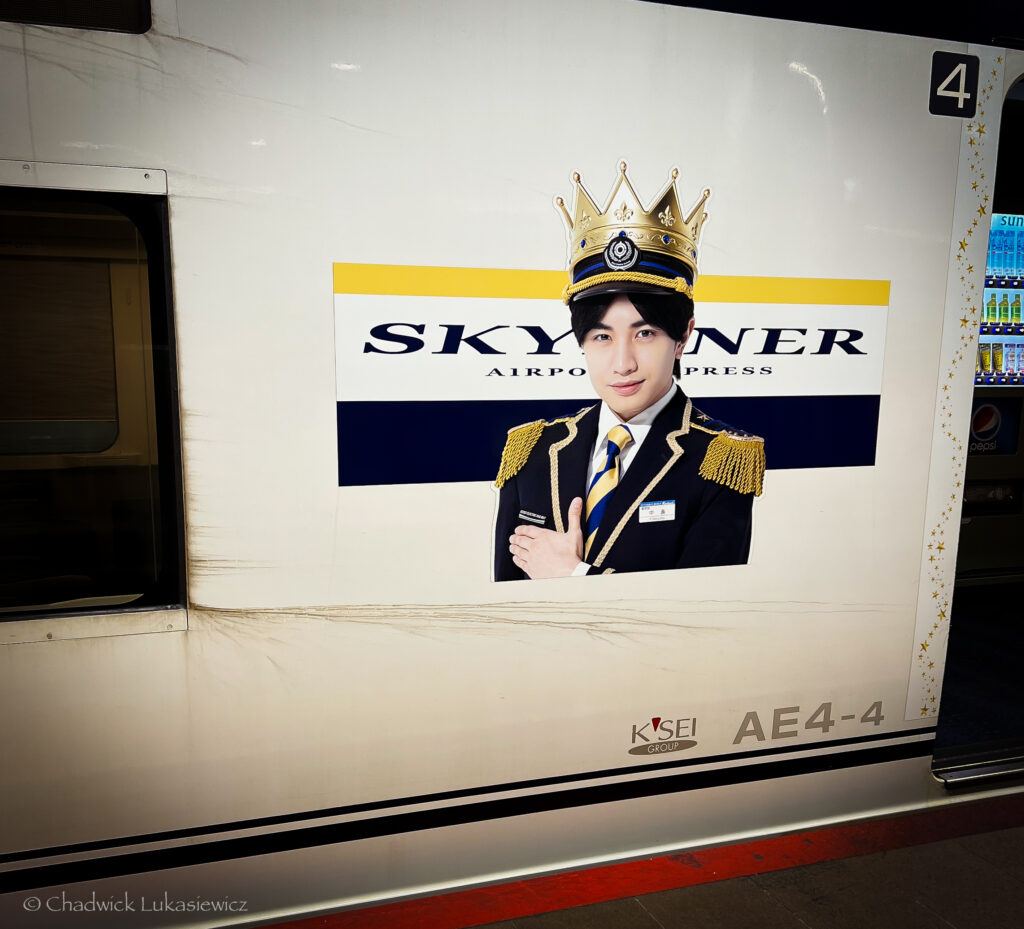 A promotional poster on the side of a Skyliner Airport Express train in Tokyo, Japan, featuring an image of a young man dressed in an elaborate uniform with gold shoulder tassels and a striped yellow-and-blue tie, embodying a regal look. He is wearing a large, ornate crown, and stands against a white background with the Skyliner branding in bold letters. The poster, located on car AE4-4, showcases the Keisei Group’s Skyliner service, adding a sense of elegance and exclusivity to the airport express train experience.