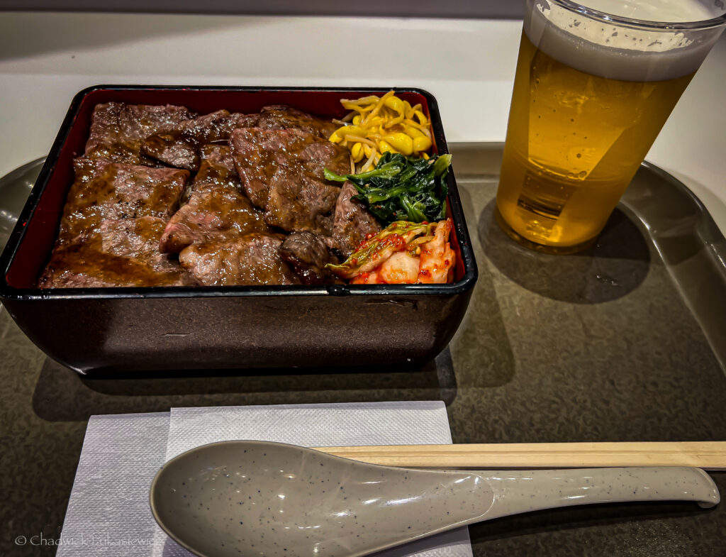 A close-up view of a hearty beef bento meal, featuring grilled beef slices, kimchi, greens, and pickled vegetables, arranged in a lacquered box. To the side, a tall glass of golden beer accompanies the meal, with chopsticks and a ceramic spoon placed on a white napkin on the tray.