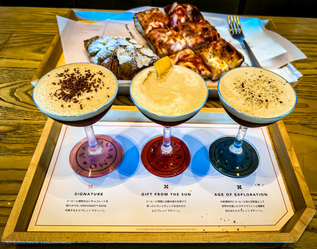 A wooden tray holds three espresso martinis in cocktail glasses, each labeled with its unique name: ‘Signature,’ ‘Gift from the Sun,’ and ‘Age of Exploration.’ Each drink has a distinct garnish, such as chocolate shavings or an orange peel. Behind the cocktails, a plate with assorted pastries, including an almond-topped croissant, sits on the table. This setup is part of the Starbucks Reserve Roastery in Tokyo, Japan.