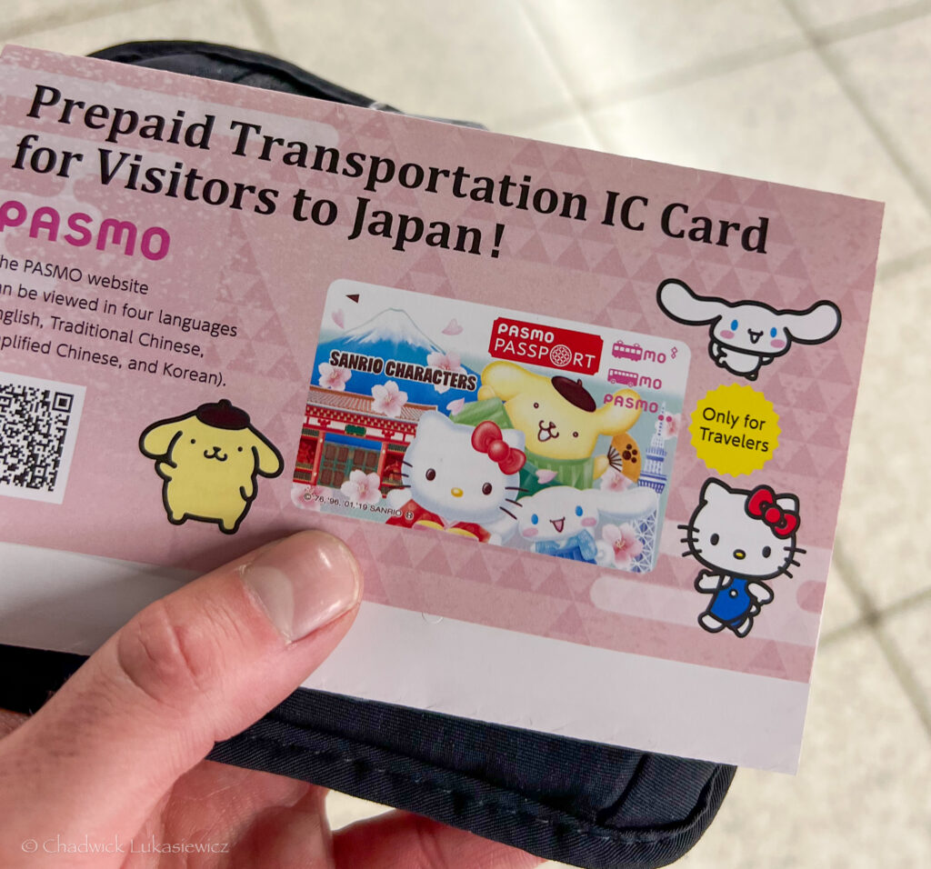 A close-up shot of a “Prepaid Transportation IC Card for Visitors to Japan” advertisement, specifically for the PASMO Passport card featuring Sanrio characters. The pink flyer displays cute illustrations of popular Sanrio characters, including Hello Kitty and Cinnamoroll, with a colorful PASMO Passport card image in the center showcasing Mount Fuji and cherry blossoms. The flyer is marked “Only for Travelers” and offers multilingual support, listing languages like English, Traditional Chinese, Simplified Chinese, and Korean. The PASMO card is ideal for tourists navigating Japan’s transportation systems