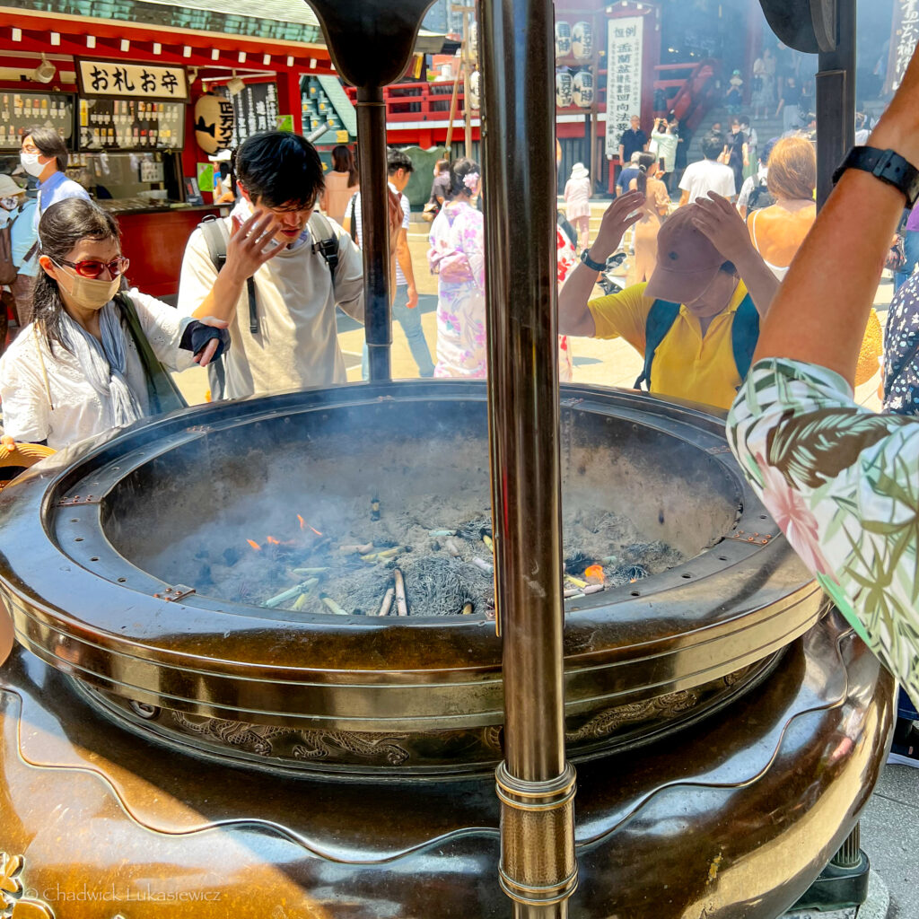 People gather around a large incense burner at Senso-ji Temple in Asakusa, Tokyo. Some individuals wave smoke toward themselves, a ritual believed to bring health and purification. The burner is filled with smoldering incense sticks, creating wafts of smoke that rise into the air. In the background, more visitors and traditional red temple structures are visible, with some people wearing summer hats and casual attire.