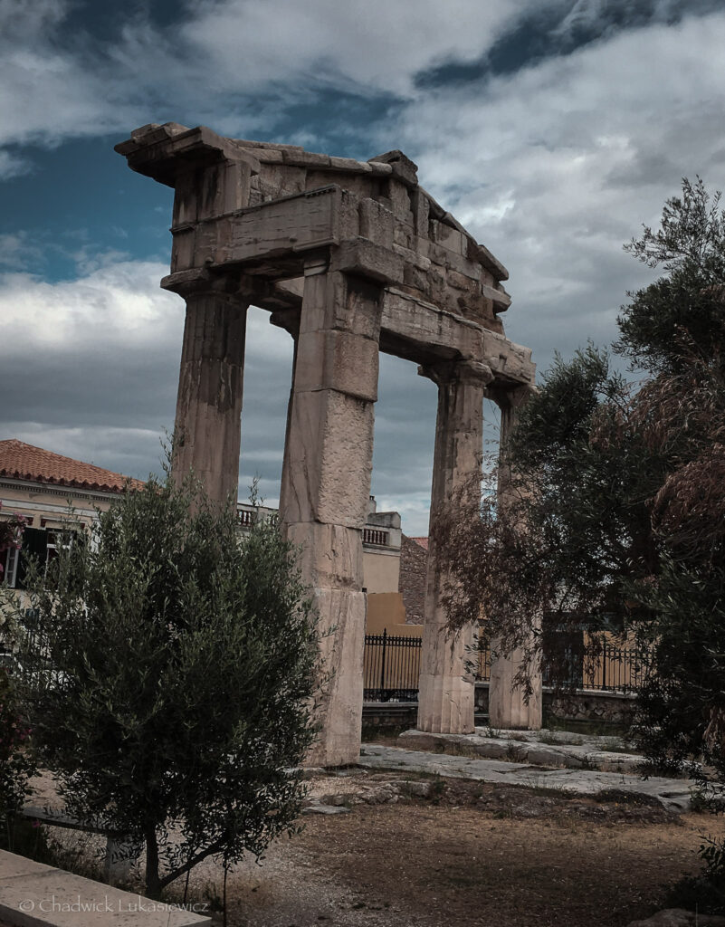 The ruins of the Roman Agora in Athens, Greece, showcasing a partially preserved stone gate with large rectangular columns and an intact architrave. The ancient structure is surrounded by olive trees and shrubs, with a backdrop of traditional houses featuring red-tiled roofs. The cloudy sky adds an atmospheric touch to the historic scene.