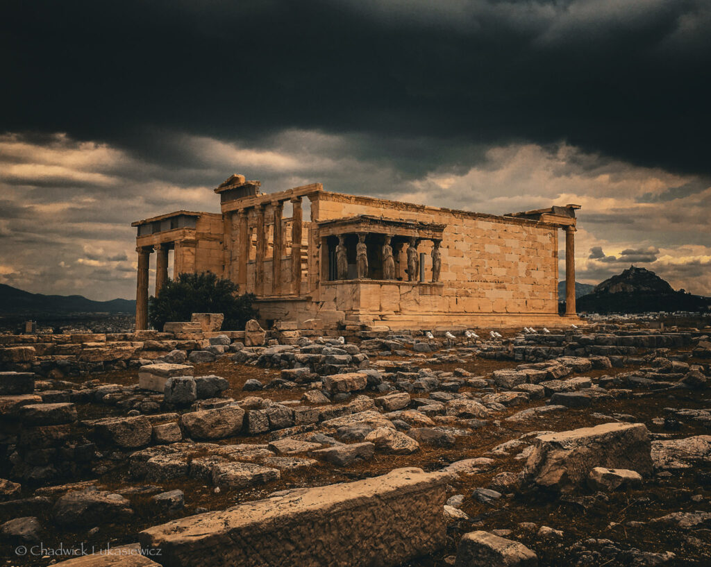 A dramatic view of the Erechtheion, an ancient temple on the Acropolis of Athens, under a stormy sky. The structure features iconic Ionic columns and the Caryatids, the sculpted female figures serving as architectural supports for the porch. The golden hues of the weathered marble contrast with the dark, foreboding clouds overhead. In the foreground, the rocky terrain is scattered with ancient stone remnants, adding to the sense of historical depth. In the distance, a hill rises on the horizon, blending with the moody and atmospheric landscape.