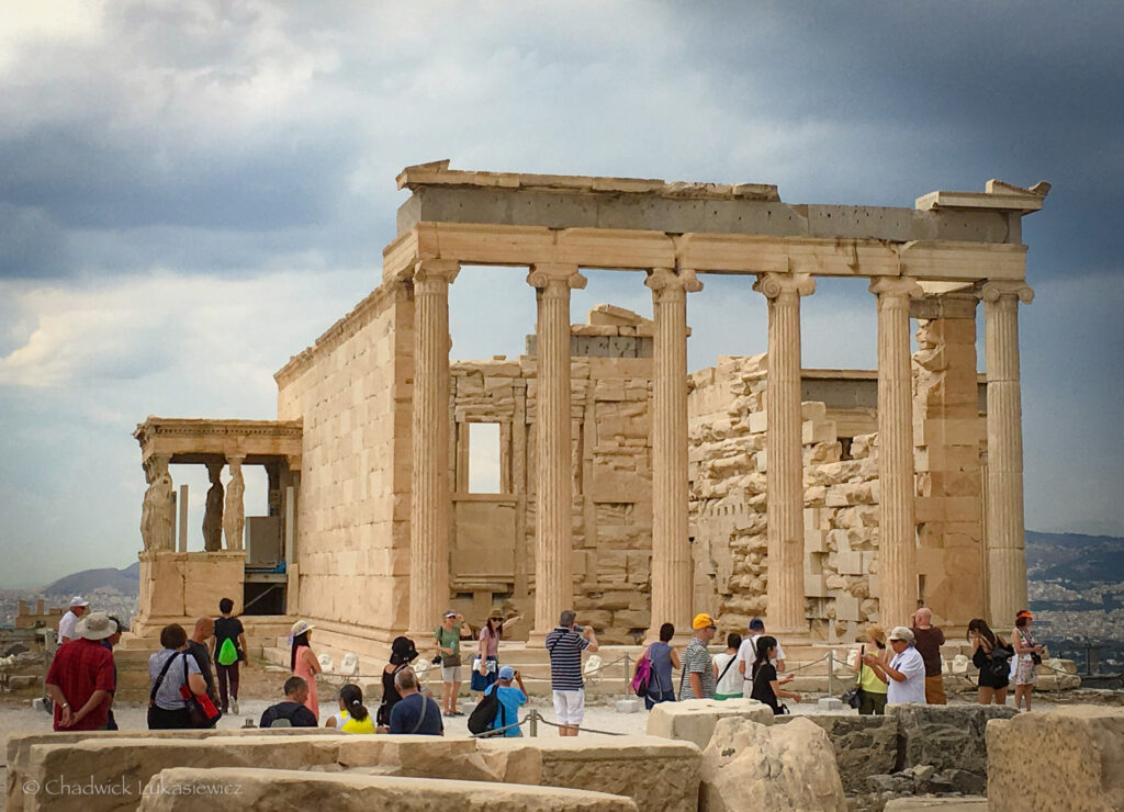 A vibrant daytime view of the Erechtheion, an ancient temple on the Acropolis of Athens, Greece. The structure features iconic Ionic columns and the famous Caryatids, sculpted female figures supporting the porch on the left. Tourists in colorful attire are gathered around the site, some taking photos and others admiring the historic architecture. The beige tones of the marble contrast with the cloudy sky, creating a dramatic backdrop. The distant cityscape of Athens is faintly visible in the background, adding depth to the scene.