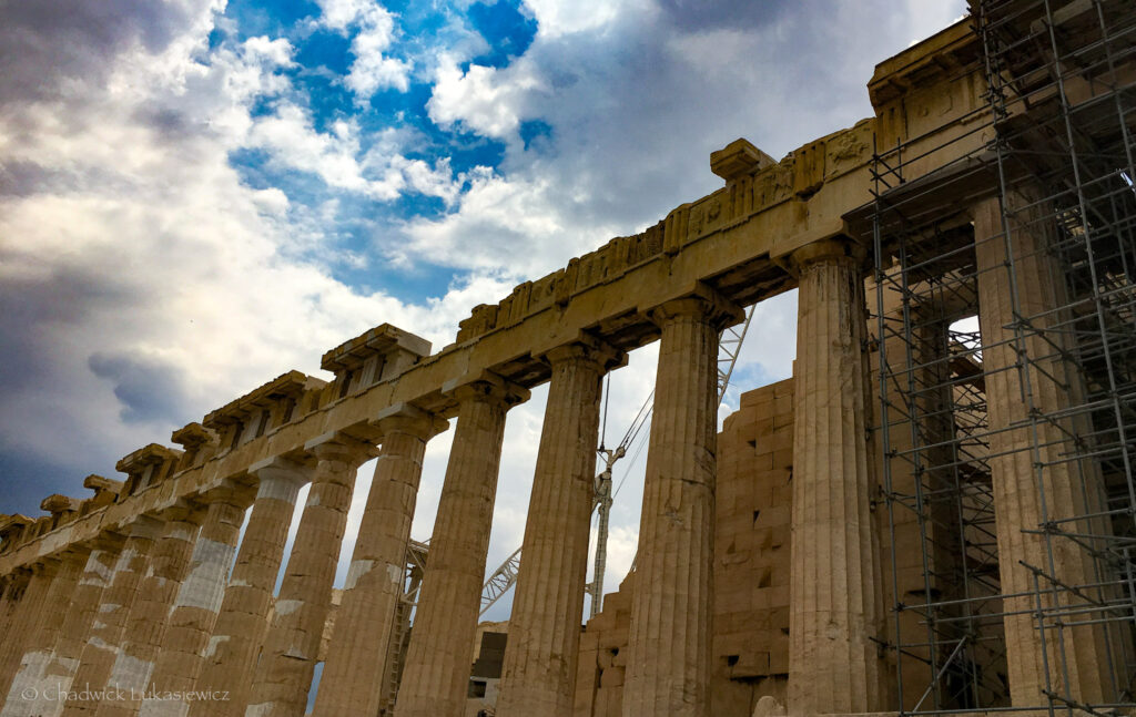 A close-up view of the Parthenon on the Acropolis of Athens, highlighting its iconic Doric columns and weathered marble. The structure is partially shrouded in scaffolding, reflecting ongoing restoration efforts. The intricate architectural details of the frieze and pediment are visible against a dynamic sky with a mix of bright blue and heavy gray clouds, adding depth and drama to the scene. The image captures the timeless grandeur of the ancient temple while acknowledging the modern preservation work being carried out to maintain its legacy.