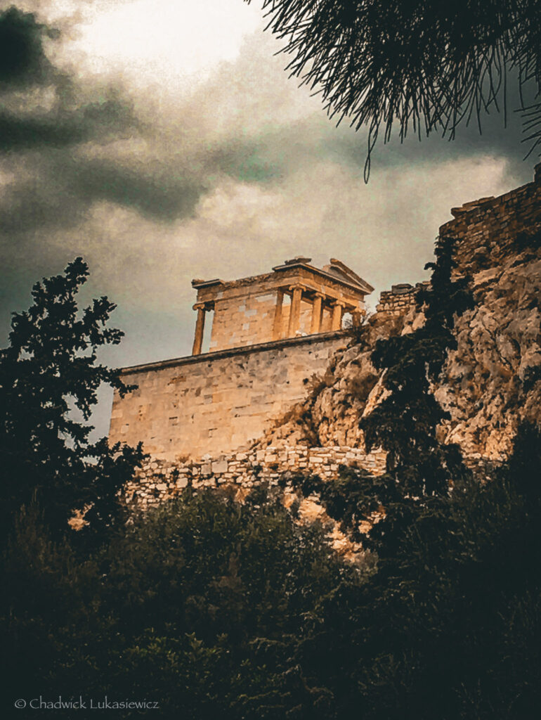 A close-up view of the Temple of Athena Nike on the Acropolis of Athens, perched atop a rocky cliff under a dramatic, overcast sky. The small yet elegant temple features Ionic columns and stands prominently against the dark green trees and rugged terrain. The moody clouds and faint sunlight create a sense of timeless grandeur.